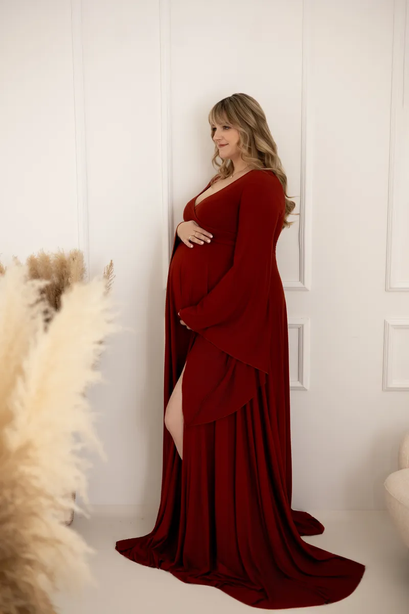 Eine schwangere Frau in einem eleganten roten Kleid posiert in einem hellen Raum.